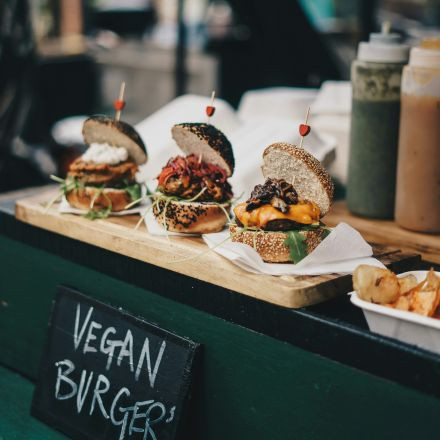 Three gourmet vegan burgers with various toppings are displayed on a wooden board at a food stall, with a sign reading Vegan Burgers.