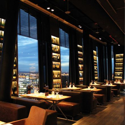 Elegant restaurant with large windows overlooking a cityscape at dusk, featuring cozy seating and shelves of wine bottles.