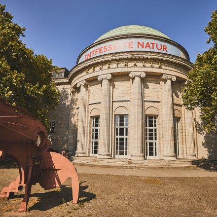 Ein neoklassizistisches Gebäude mit Säulen und einer Kuppel, auf dem Schild steht „Entfesselte Natur“, davor eine rote Metallskulptur.