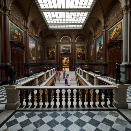 Eine große, reich verzierte Halle mit Schachbrettmusterboden, großen Gemälden, Statuen und Menschen, die unter einer Glasdecke neben einer Treppe entlanggehen.