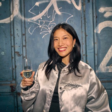 Frau in silberner Jacke hält ein Glas Weißwein und lächelt vor einer blauen Wand mit Graffiti.