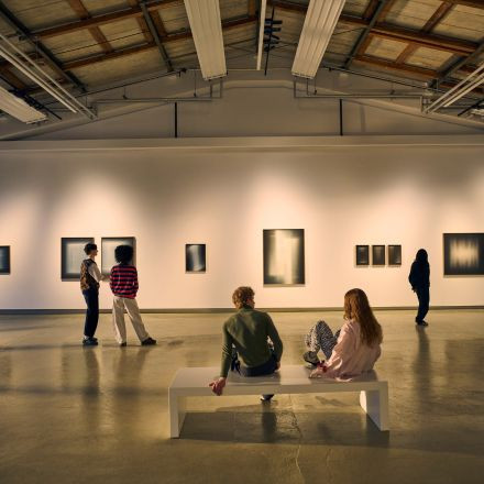 Menschen betrachten abstrakte Kunst an einer Galeriewand; zwei sitzen auf einer weißen Bank, während andere stehen und die Kunstwerke betrachten.