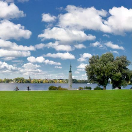 Eine grüne Wiese an einem See mit zwei Radfahrern, einer Statue, Bäumen und einem blauen Himmel mit vereinzelten weißen Wolken.