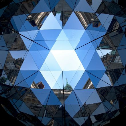 Geometrisch verspiegelte Decke, die den blauen Himmel, Gebäude und die Stadtlandschaft reflektiert und mit dreieckigen Glasplatten ein sechseckiges Muster bildet.