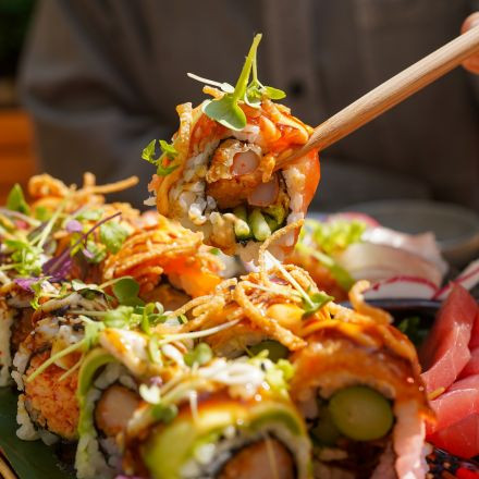 A close-up of colorful sushi rolls topped with microgreens, with chopsticks holding one piece above the plate.