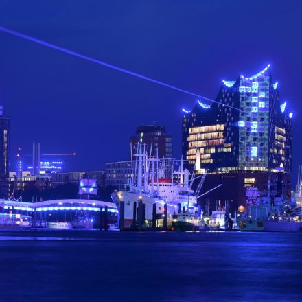 Beleuchtetes modernes Gebäude und Schiffe auf einem Fluss bei Nacht, leuchtend blaue Lichter spiegeln sich im Wasser.