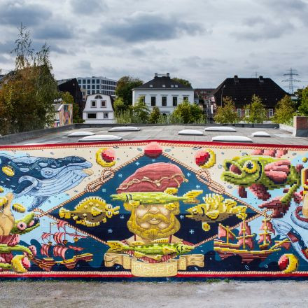 Farbenfrohes Wandgemälde mit Meereslebewesen, Schiffen und einer bärtigen Figur in der Mitte, vor Häusern und bewölktem Himmel im Hintergrund.