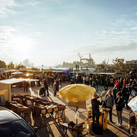 A busy outdoor market with people browsing stalls under a sunny sky, with ships and cranes visible in the background.