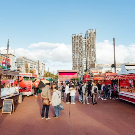 People stroll and shop at an open-air market with colorful stalls, modern buildings, and a blue sky in the background.