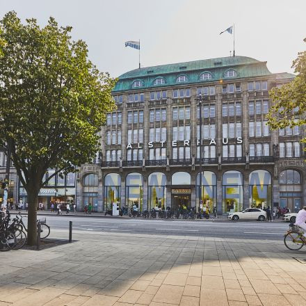 Ein großes Gebäude mit „ALSTERHAUS“ an der Fassade, davor Bäume und Fahrräder und Menschen, die die Straße entlanggehen.