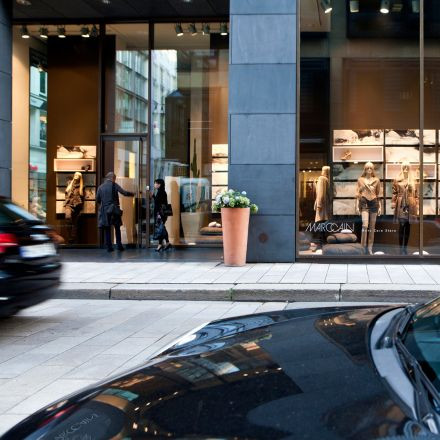 Zwei Personen betreten ein modernes Bekleidungsgeschäft mit großen Glasfenstern, in denen Schaufensterpuppen und Regale ausgestellt sind, davor parken Autos.