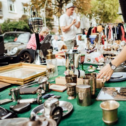 A person browses for vintage items on a table at an outdoor flea market, with people and trees in the background.