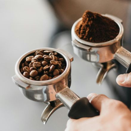 Two portafilters: one filled with whole coffee beans, the other with ground coffee, held by a person.