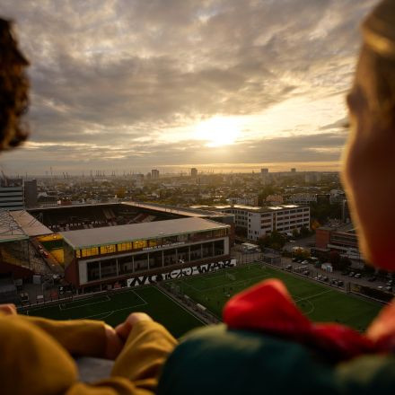 Zwei Personen blicken bei Sonnenuntergang auf eine Stadt und ein Fußballfeld, mit Gebäuden und einem dramatischen Himmel im Hintergrund.