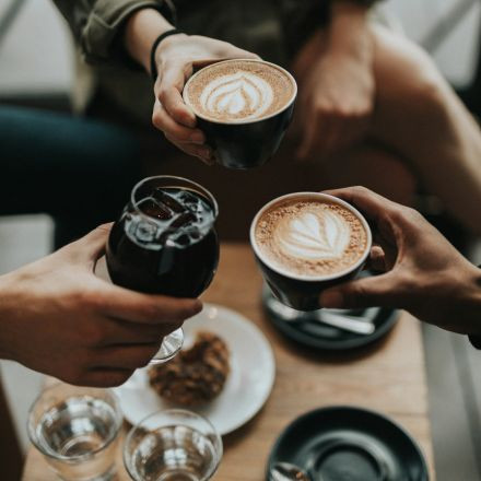 Three people toast with two cups of latte art coffee and a glass of iced drink on a table with water and a cookie.