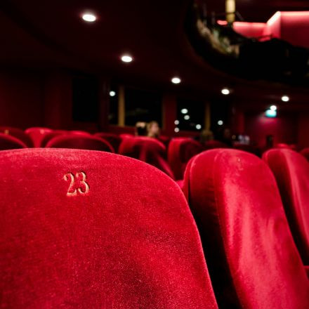 Close-up of a red velvet theater seat numbered 23, with other red seats and the dimly lit theater in the background.