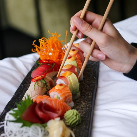 Eine Hand nimmt mit Stäbchen ein Stück buntes Sushi von einem Tablett mit verschiedenen Sashimi und Beilagen.