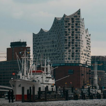An einem bewölkten Tag steht hinter einem angedockten weißen Schiff ein modernes Glasgebäude mit gewelltem Dach.