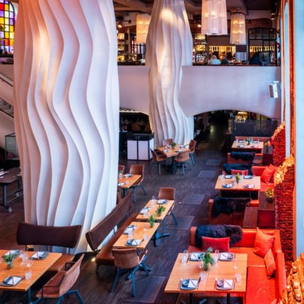 Modernes Restaurant-Interieur mit hohen, gewellten weißen Säulen, Holztischen, gemütlichen Sitzgelegenheiten und warmer Beleuchtung.