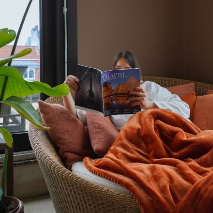 Eine in eine orangefarbene Decke gehüllte Person liest ein Reisemagazin, während sie auf einem gemütlichen Sofa in der Nähe eines Fensters mit Pflanzen in der Nähe entspannt.