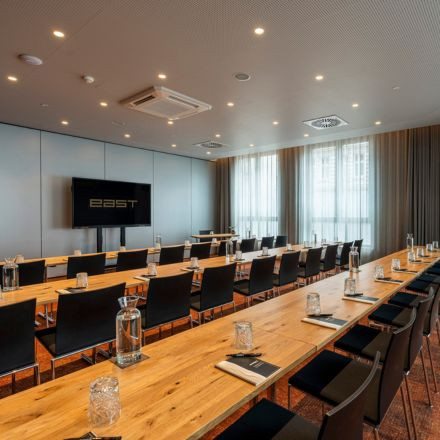 Moderner Konferenzraum mit Holztischen, schwarzen Stühlen, Glaswasserflaschen, Notizblöcken und einem großen Bildschirm davor.
