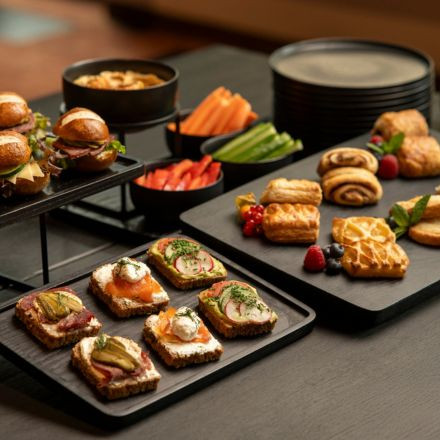 Eine Auswahl an Sandwiches, Gebäck und frischem Gemüse, angerichtet auf schwarzen Tabletts und Tellern auf einem dunklen Tisch.