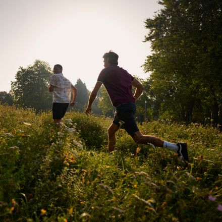 Zwei Menschen laufen unter einem hellen, sonnigen Himmel durch eine von Bäumen umgebene Wiese mit Wildblumen.