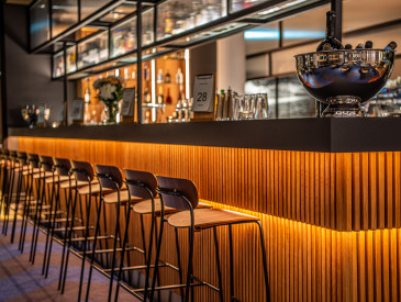 Moderne Bar mit Holzhockern, orange beleuchtetem Tresen und Regalen voller Flaschen und Gläser