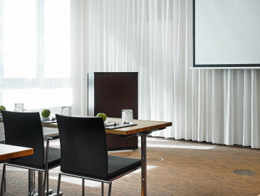 Modern conference room with tables, black chairs, glasses, green apples, notepads, and a projection screen in front of white curtains.
