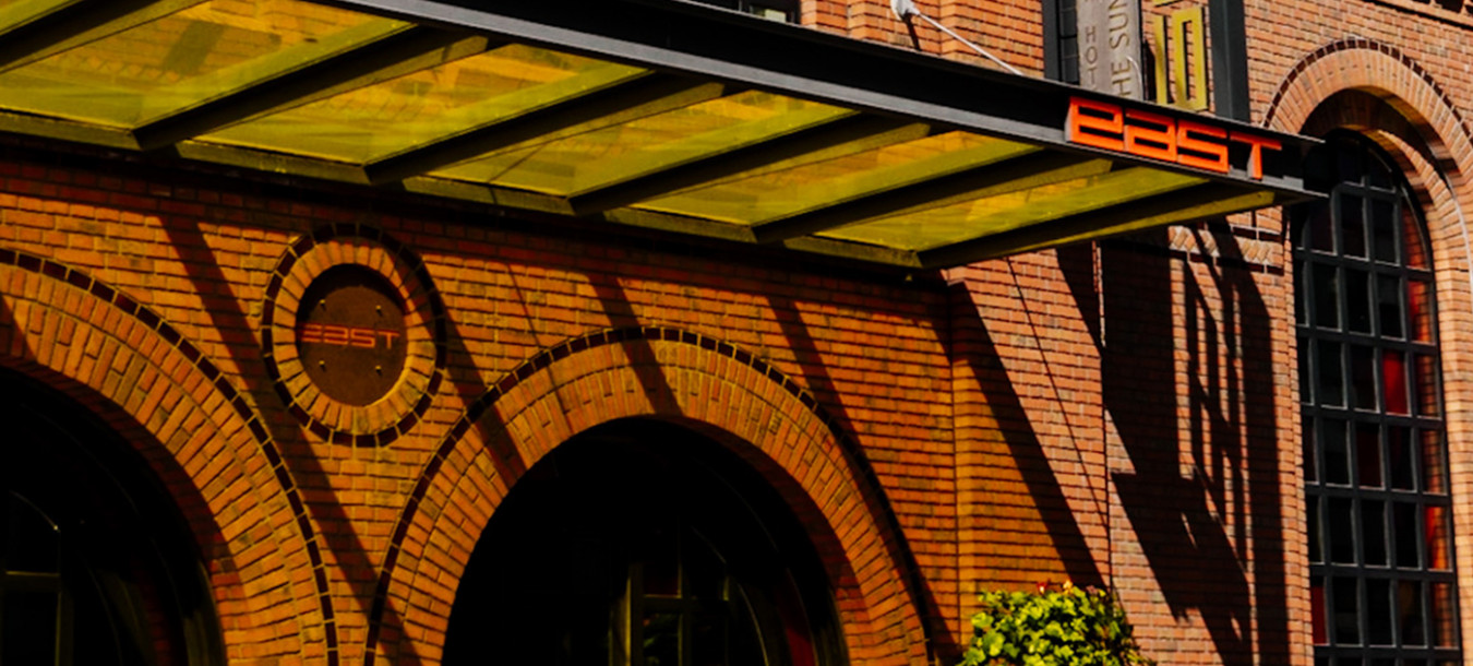 Sunbeams fall on the brick facade of the east Hotel, which features large arched windows and a glass-enclosed upper floor, a covered entrance, a large black and gold sign, and a potted plant next to the door.