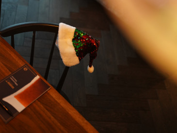 Eine paillettenbesetzte Weihnachtsmütze hängt auf einem schwarzen Stuhl neben einem Holztisch in einem gemütlichen, schwach beleuchteten Raum mit einem Bett in der Nähe.