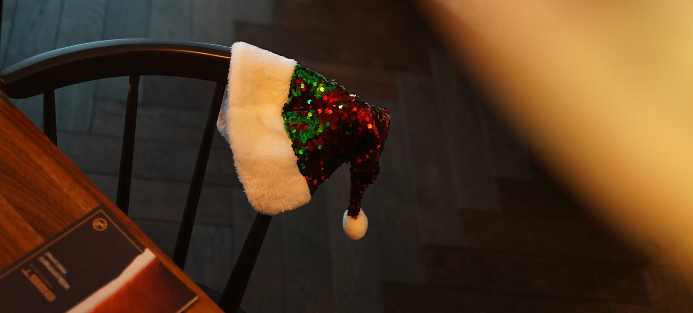 Eine paillettenbesetzte Weihnachtsmütze hängt auf einem schwarzen Stuhl neben einem Holztisch in einem gemütlichen, schwach beleuchteten Raum mit einem Bett in der Nähe.