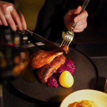 Eine Person schneidet in einem Restaurant eine gebratene Ente an, die mit Rotkohl und einer kleinen Kartoffel auf einem dunklen Teller serviert wird.
