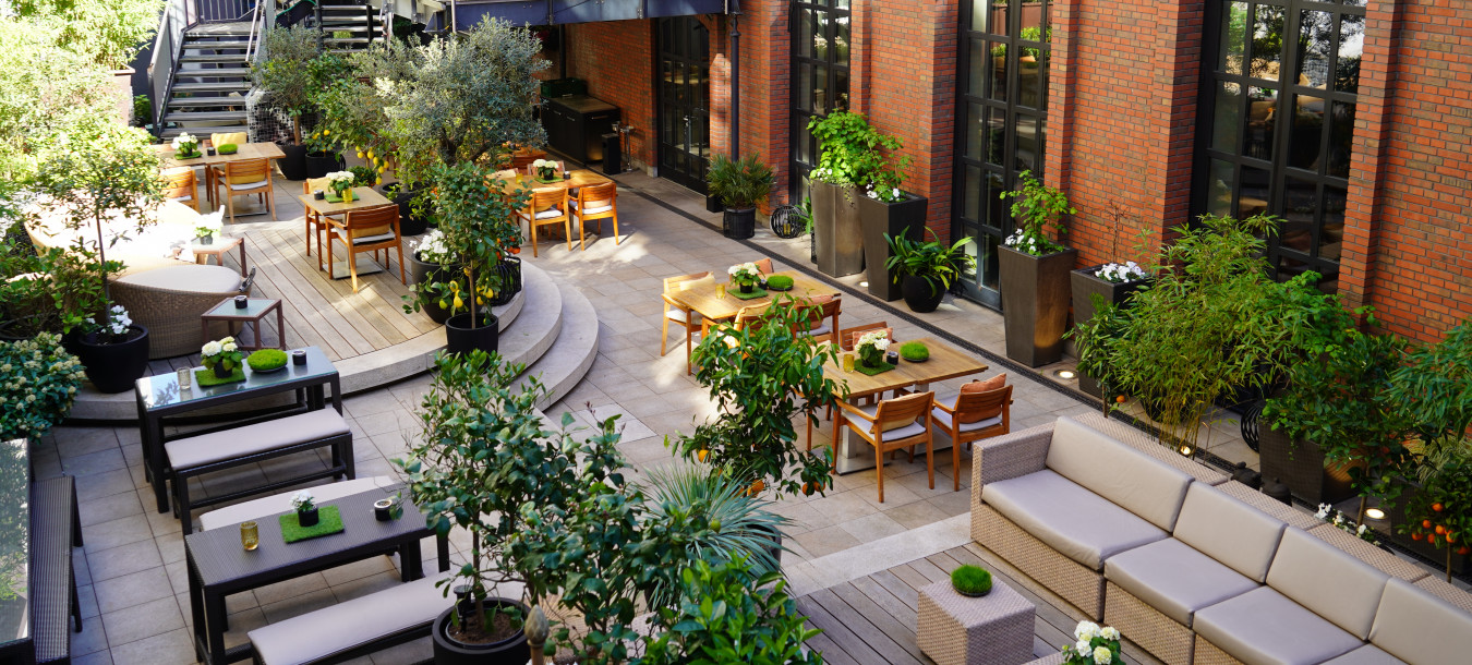 A modern outdoor patio with wooden tables, chairs, sofas, lush plants, and an upper deck with a large white umbrella.