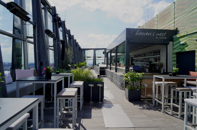 Rooftop bar with modern tables, stools, plants, and a central bar under a partly cloudy sky.