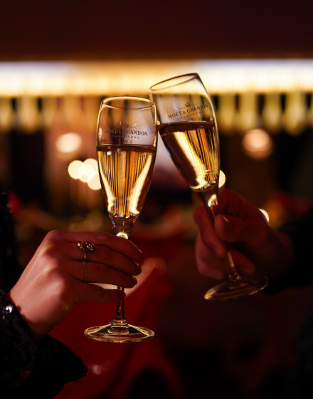 Zwei Hände, die in einer schummrigen Umgebung mit verschwommenen Lichtern im Hintergrund mit Moët & Chandon-Champagnergläsern anstoßen.