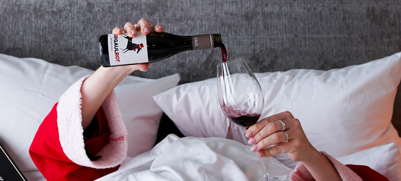 Eine Person in einem roten Gewand gießt Rotwein aus einer kleinen Flasche in ein Weinglas, während sie in einem Bett mit weißen Kissen und grauem Kopfteil liegt.