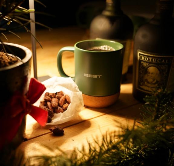 Eine grüne Tasse, eine Tüte Nüsse und zwei dunkle Flaschen auf einem Holztisch, umgeben von festlichem Grün und einer Pflanze mit roter Schleife.