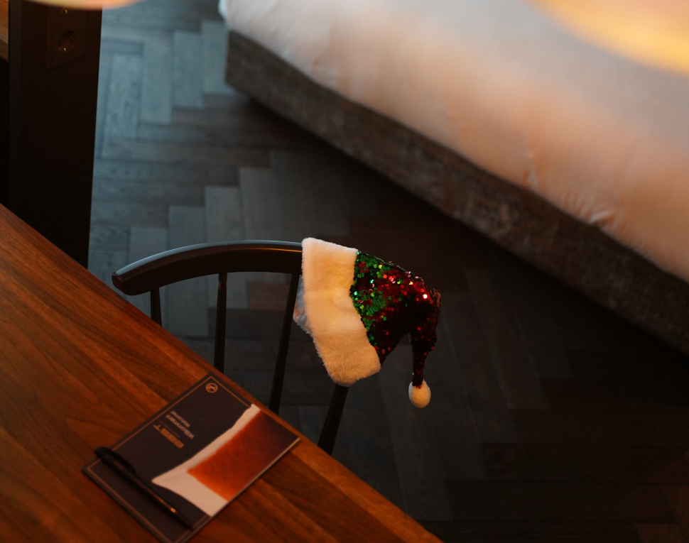 Gemütliches Zimmer mit warmer Beleuchtung, Holztisch und einem schwarzen Stuhl, der mit einer glitzernden Weihnachtsmütze geschmückt ist. In der Nähe steht ein einladendes Bett in weicher Unschärfe.