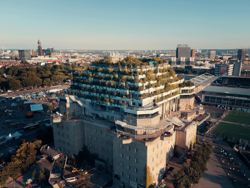 Luftaufnahme eines großen, stufenförmigen Gebäudes, bedeckt mit Grün und Bäumen, umgeben von der Stadtlandschaft und einem nahe gelegenen Sportplatz.