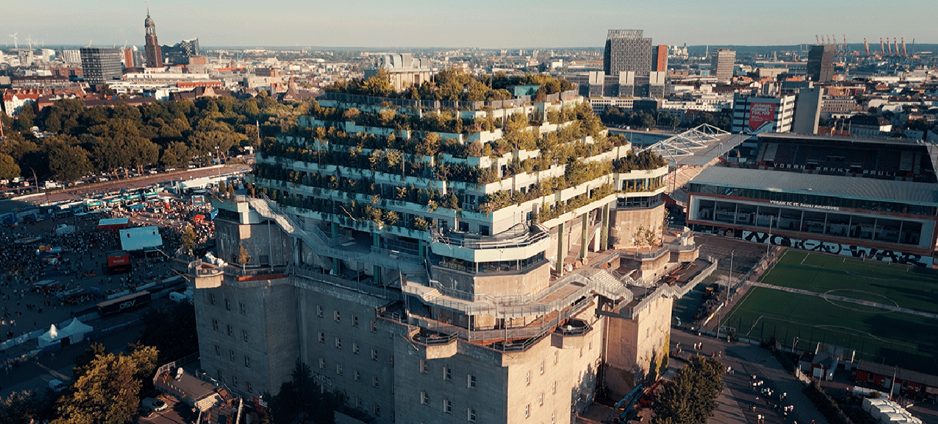 Ein moderner Stufenbau mit begrünten Dachterrassen steht in einer Stadtlandschaft, umgeben von Bäumen und urbanen Strukturen.