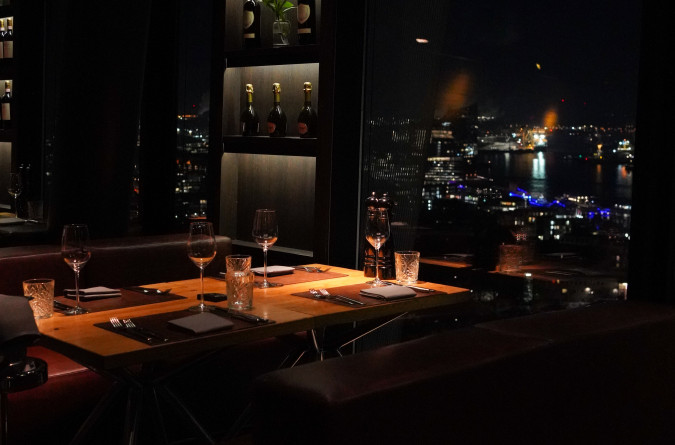 Von einem schwach beleuchteten Restauranttisch am Fenster hat man Ausblick auf eine Stadtlandschaft, deren Lichter sich nachts auf dem Wasser spiegeln.