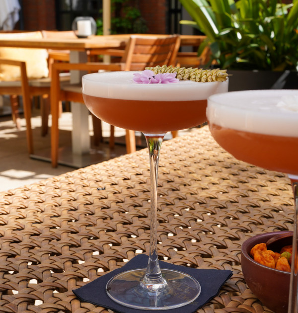 Ein mit Blumen und Früchten garnierter Cocktail steht auf einem geflochtenen Tisch auf einer Außenterrasse mit Stühlen und grünen Pflanzen im Hintergrund.