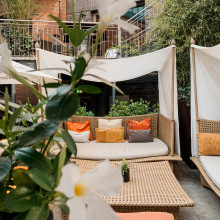 Außenterrasse mit geflochtenen Loungebetten, bunten Kissen, weißem Baldachin und grünen Pflanzen; im Vordergrund eine weiße Blume.