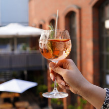 Eine Hand hält ein Glas rosa Cocktail mit Garnitur, im Freien in der Nähe eines Backsteingebäudes mit großen Fenstern und Sonnenschirmen.