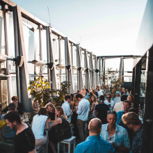 Eine Gruppe von Menschen unterhält sich bei Tageslicht in einer Bar auf dem Dach, umgeben von modernen Glaspaneelen und Stadtblick.
