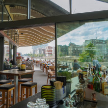Modernes Restaurant am Flussufer mit Holzdecke, Glaswänden, Tischen, Barbereich und an einem sonnigen Tag von draußen sichtbaren Stadtgebäuden.