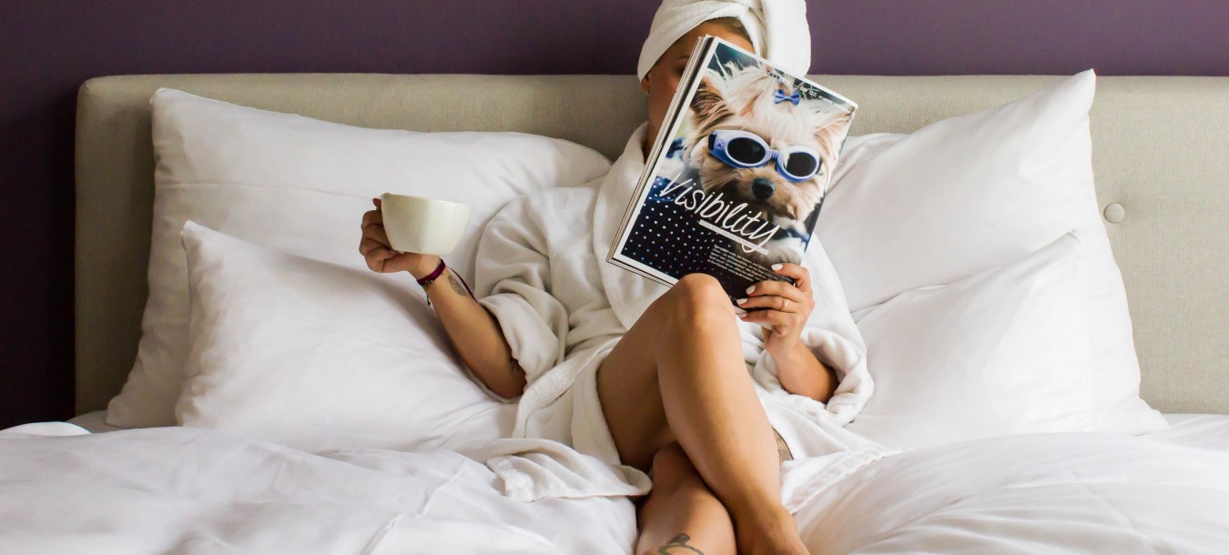 Frau sitzt mit Kaffee und Zeitung im Bademantel auf dem Bett.