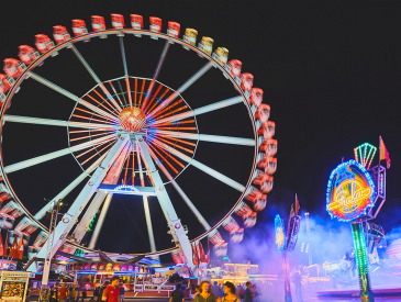 Bei einem lebhaften Jahrmarkt dreht sich nachts ein hell erleuchtetes Riesenrad, im Vordergrund sind bunte Lichter und Menschen zu sehen.