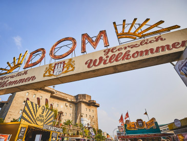 Entrance archway to Hamburg DOM fair with Herzlich Willkommen and decorative lights, festival stalls, and blue sky in the background.